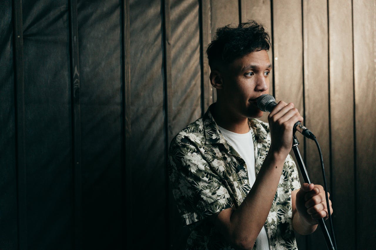 A young male singer in a floral shirt performing indoors with a microphone.