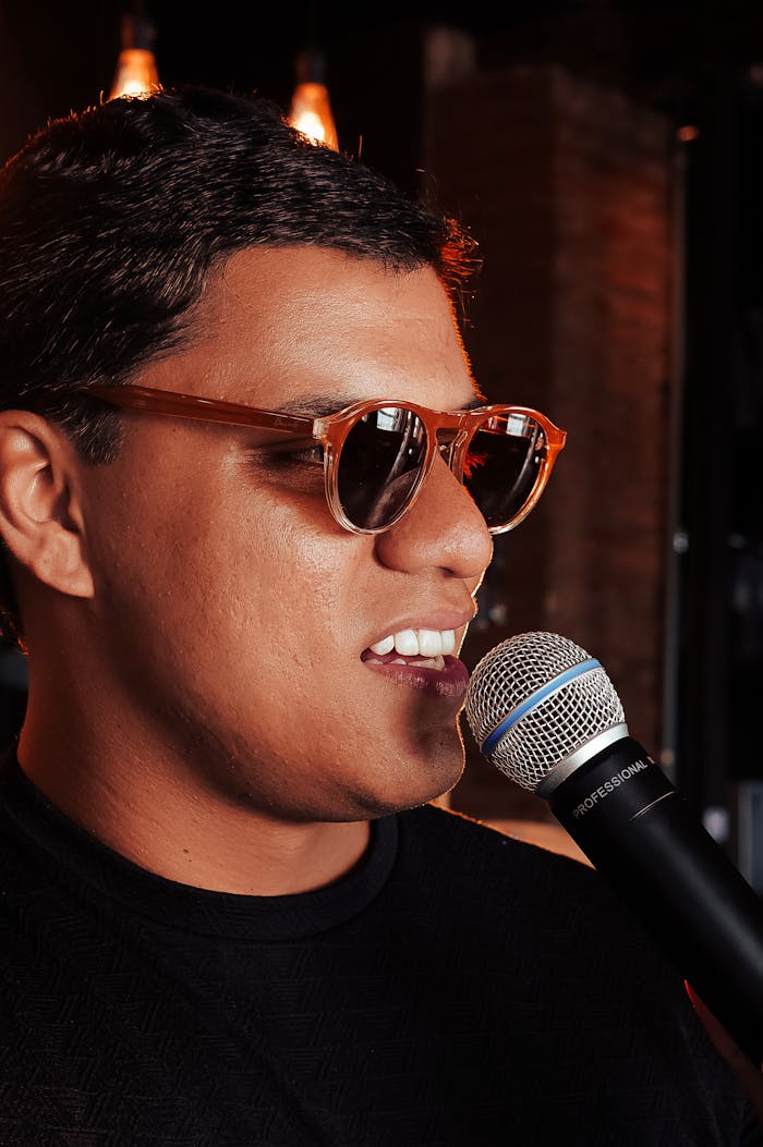Young man in sunglasses singing passionately into a microphone indoors with ambient lighting.